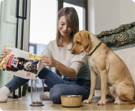 Γυναίκα που ρίχνει τροφή της Hill's Pet Nutrition σε ένα δοσομετρικό δοχείο, ενώ ένας σκύλος την παρακολουθεί με προσοχή.
